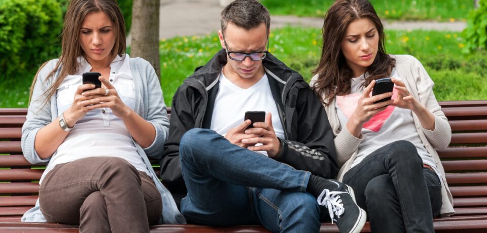 Group,Of,Friends,Two,Women,And,One,Man,,Sitting,On three people on bench looking down at cell phone with tech neck